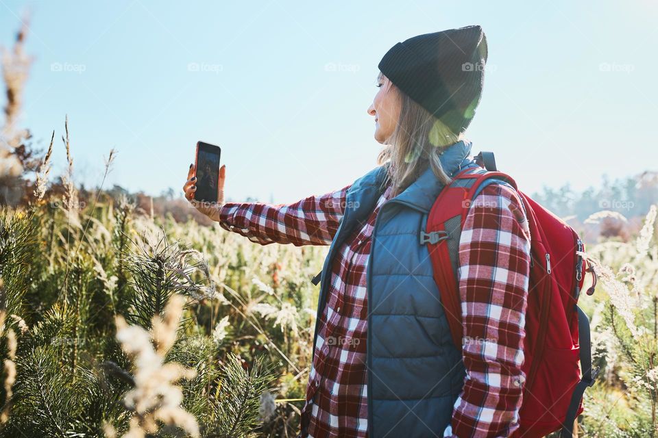 Young woman taking selfie with smartphone during vacation trip in mountains. Woman with backpack hiking through tall grass along path on meadow. Spending summer vacation close to nature