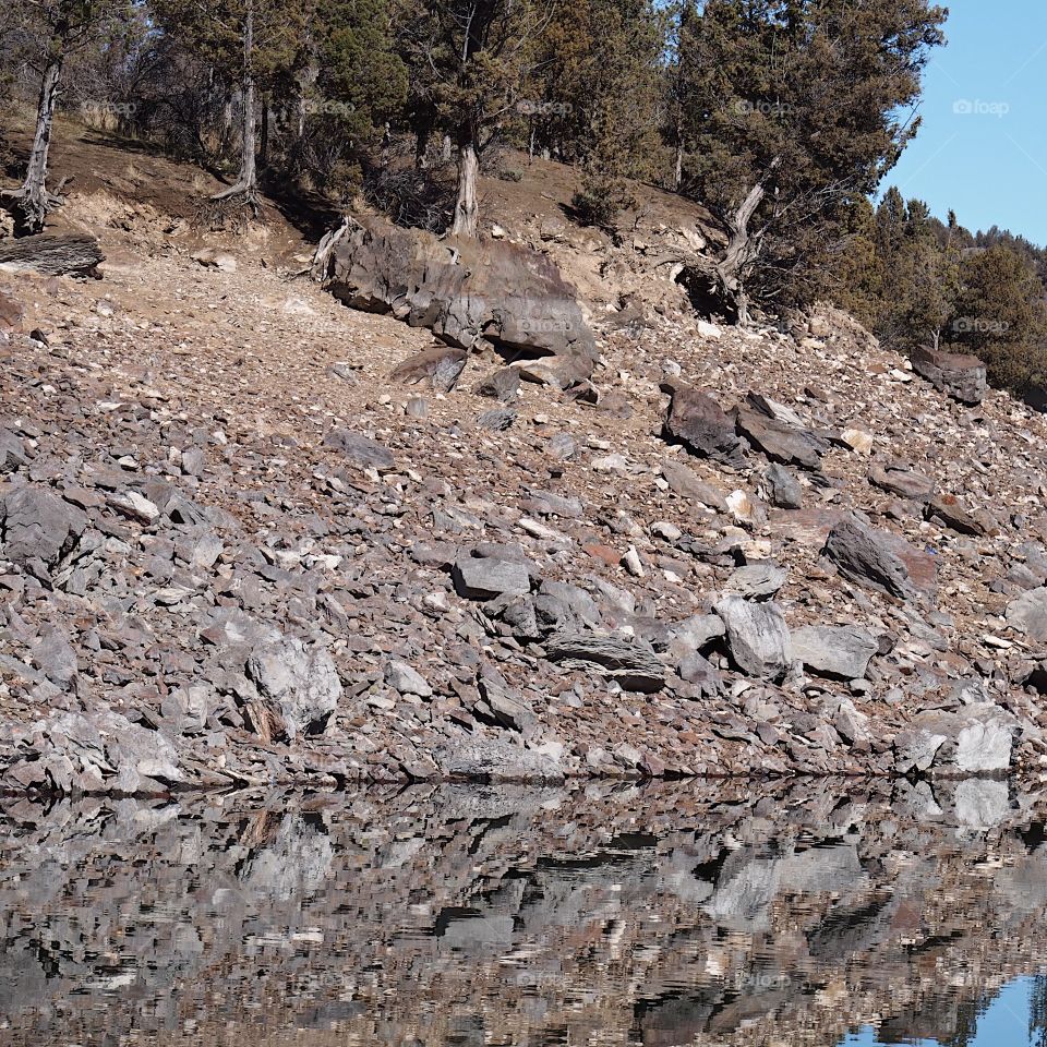 Trees and rocks on the steep slopes of the shores of Ochoco Reservoir in Central Oregon reflect in the glasslike waters on a beautiful sunny spring day with clear blue skies. 