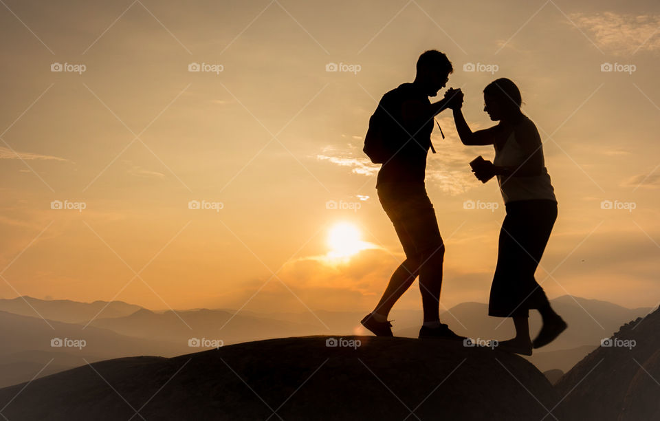 Silhouette of a man helps woman walking on the rocks in front of the sunset.