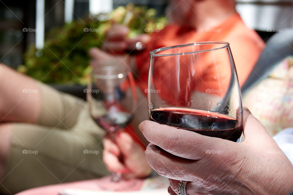 Three people holding three wine glasses filled with red wine on a porch in the sunlight. The foreground glass is in sharp focus with an older woman’s hand holding it.