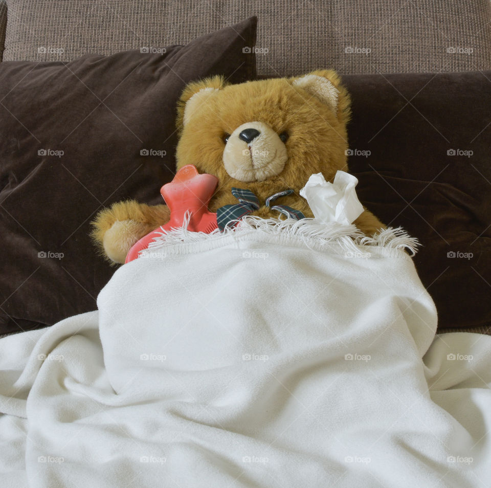 the poor teddy has got a cold. he is lying on the couch with a hotwaterbottle
