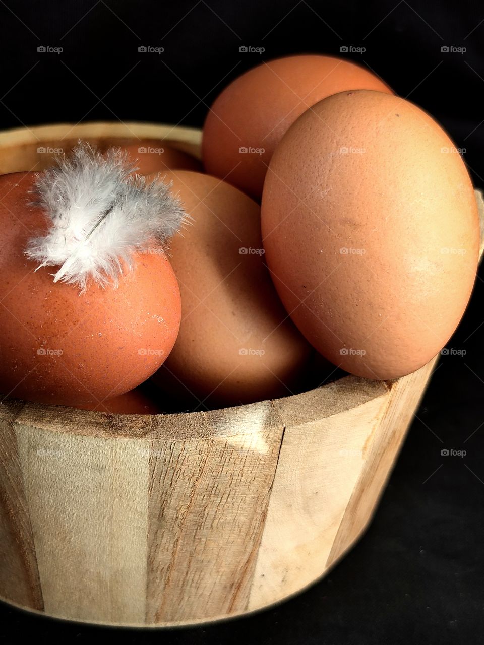 a wooden box containing brown chicken eggs.  a chicken feather lies on one egg.  Black background