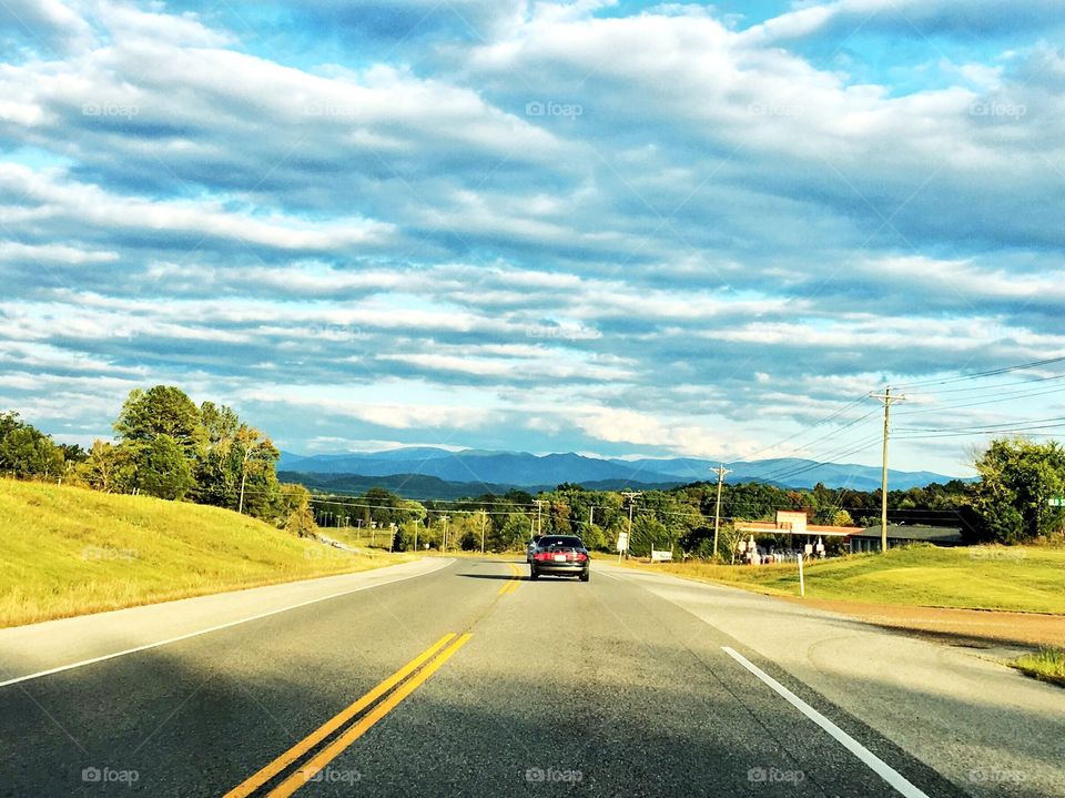 A country highway on a bright, cloudy day. One car can be seen driving into the background of blue mountain ridges. 