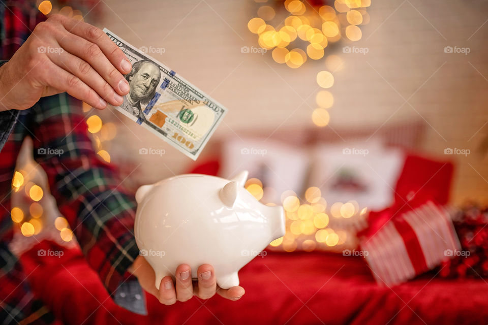 piggy bank with 100 dollars banknote in festive New Year atmosphere of scenery.  Magical bokeh with Christmas tree and bright lights.