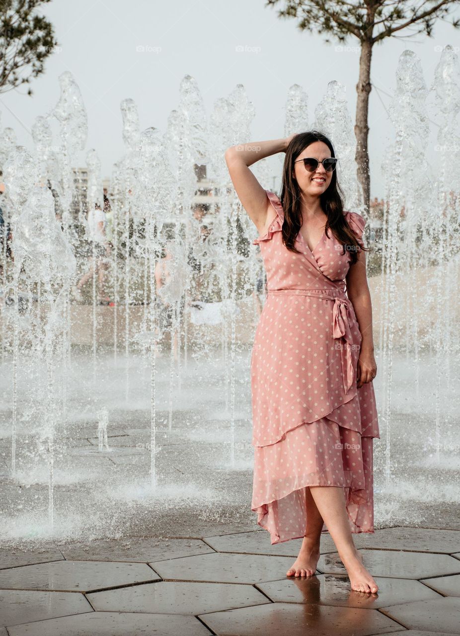 Full length photo of young woman in pink dress standing in front of fountain in city