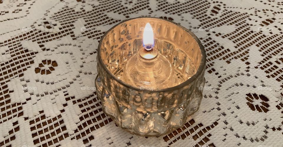 Oil light in glass jar within a metal votive holder on a white lace doilie