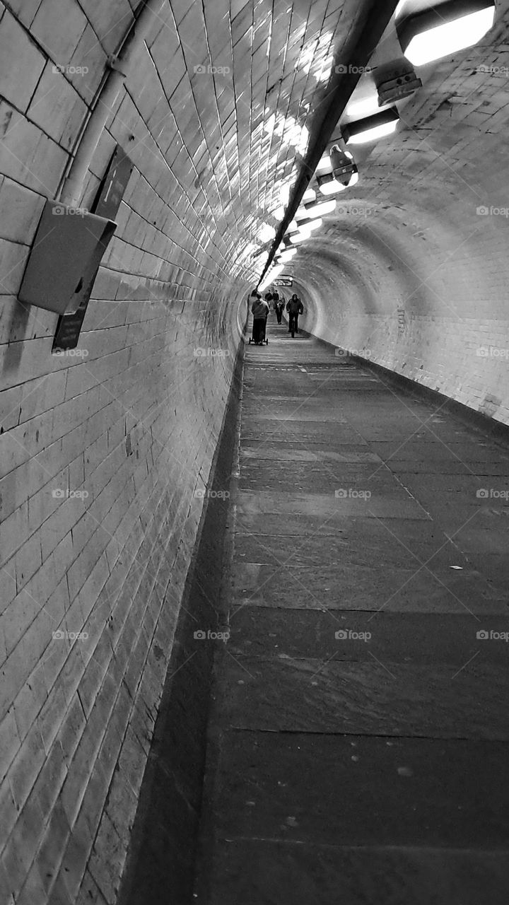 Bkack and white image of an underground foot tunnel. Cylindrical walls and floor. people in background.