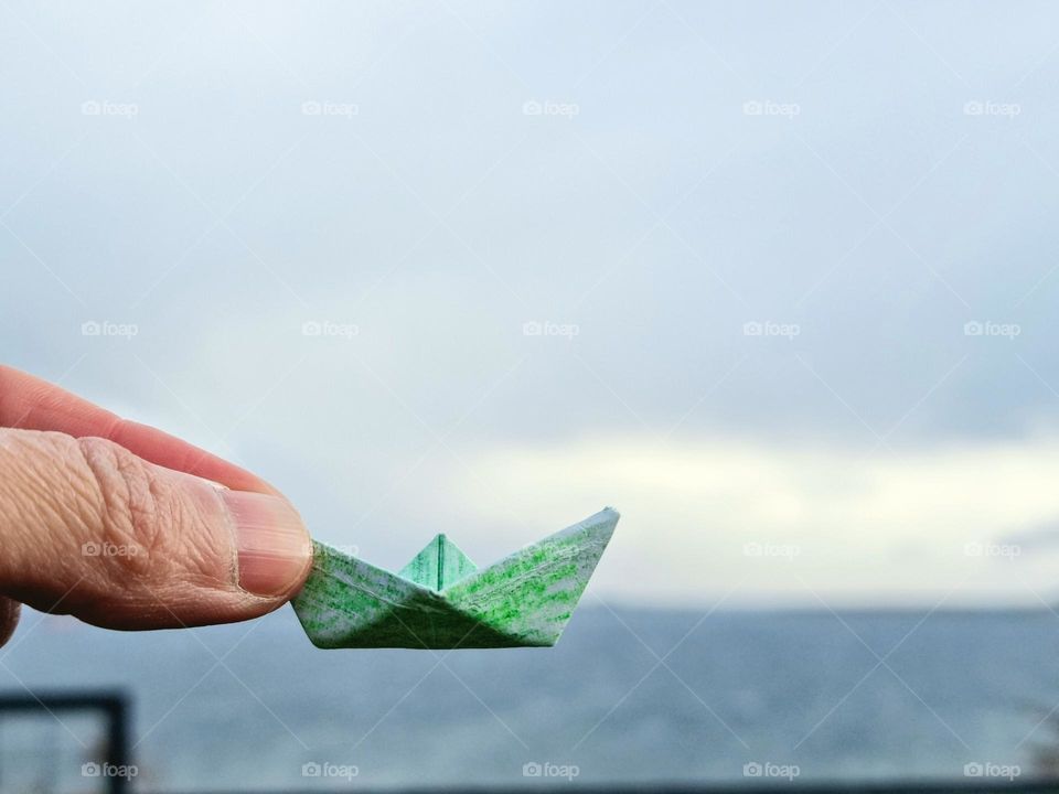 a green paper boat held by one hand, in the background and out of focus the sea and the sky.