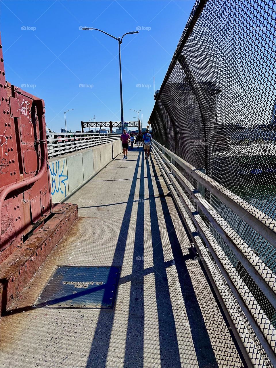 These dramatic guideline shadows are cast onto the pedestrian lane of the “Pulaski Bridge” at “Newtown Creek” that connects LIC to “Greenpoint”, Brooklyn, the direction that we are facing by the late afternoon sun. 2024. Hypnotic Productions