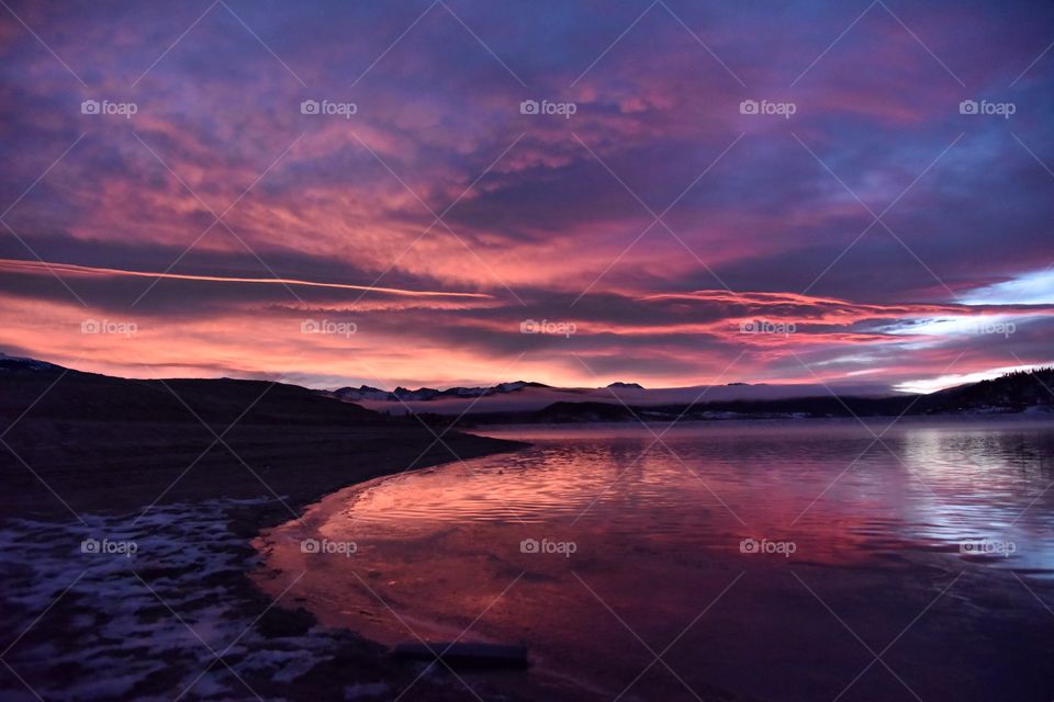 A sunrise on a cold winter morning in the mountains. The scene is set with snow and pine trees. The reflection induces awe and wonder. Pink is vivid and clouds are deep.