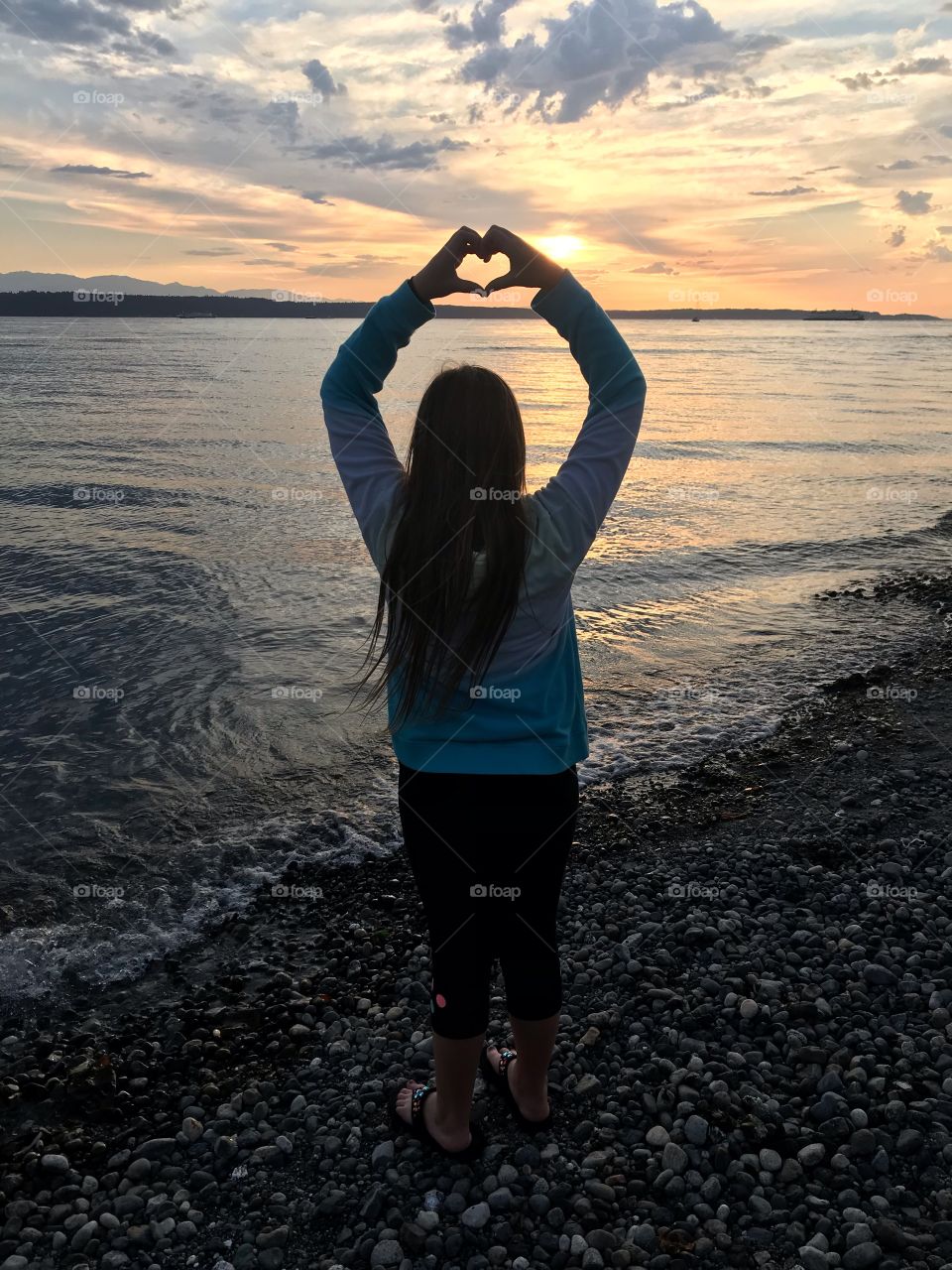 Gorgeous photo of young girl putting her hands together to form a heart with beautiful sunset in front of her! 