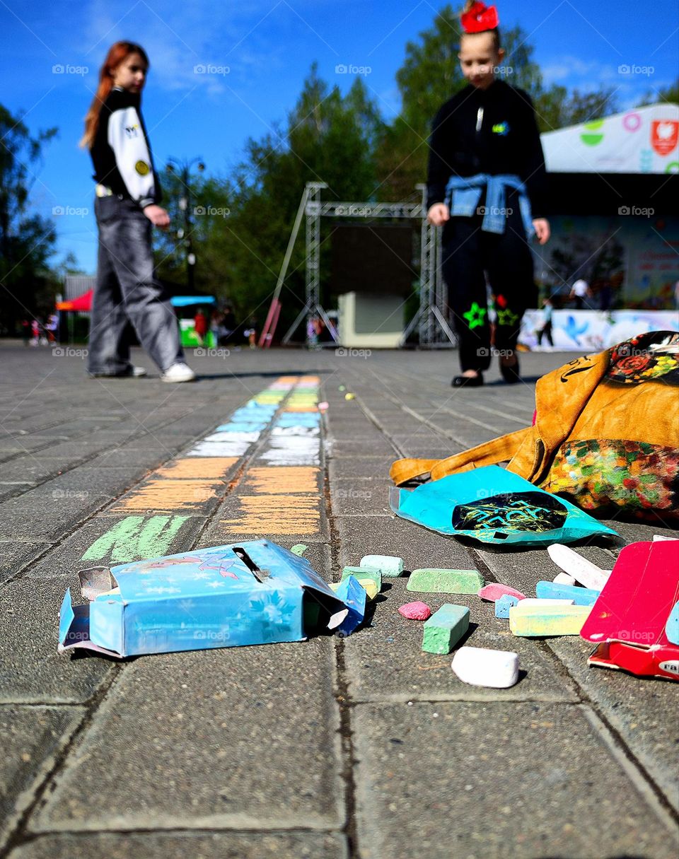 View above the ground.  Asphalt tiles with colorful crayons, boxes and a bag.  In the background are two girls who drew with chalk on the asphalt tiles.