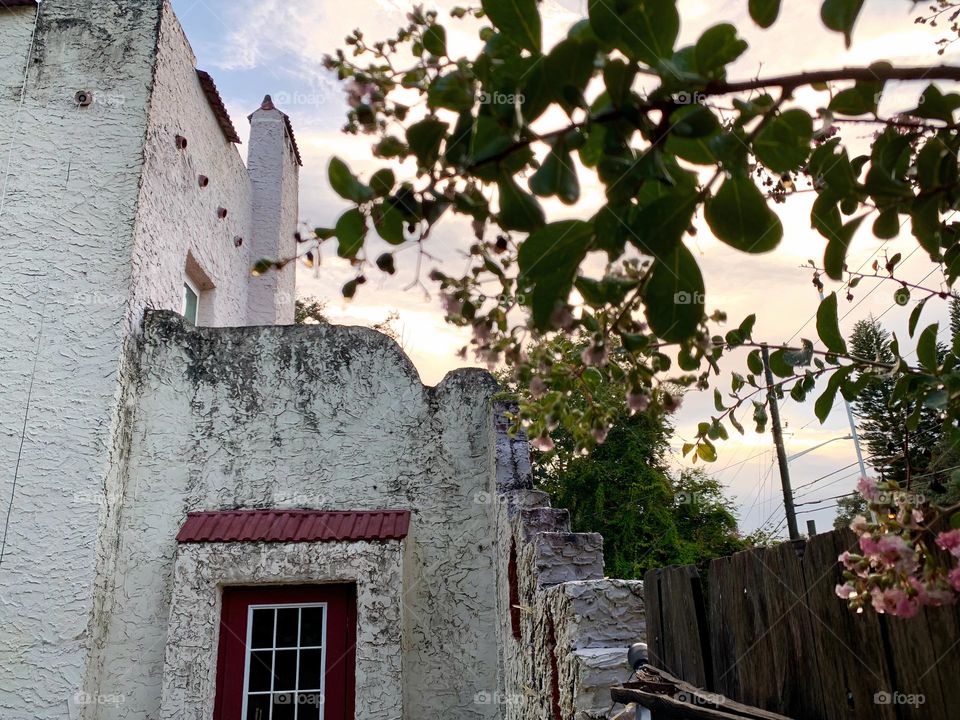 White and red spanish old style architecture residential large house built in the early 1900s. Dark wine red door with window on the side with aging signs focusing on tree branch with wet flowers and leaves by stairs style wall and wooden door.