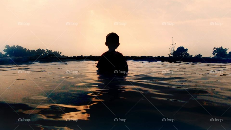 a little boy spending time in nature pool,  under the shimmering sunlight of yellow sunset..