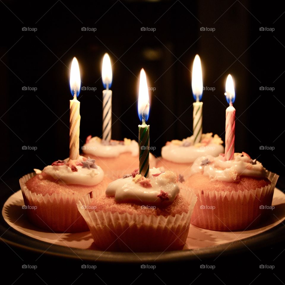 Five iced cupcakes on a plate with lit birthday candles in the dark. 