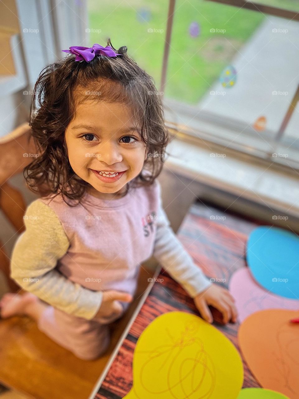 Toddler girl smiles while making Easter decorations for the home, making homemade Easter decor, making rabbit heads for the home, crafting rabbits out of colorful paper, making rabbits for Easter with a toddler girl