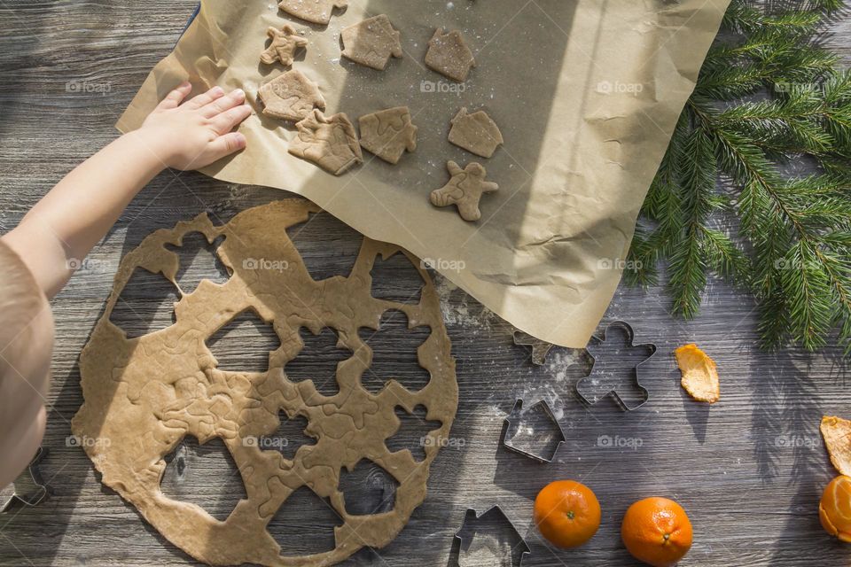 Little child helps cooking christmas ginger cookies on a wooden table with tangerines and green Christmas trees.