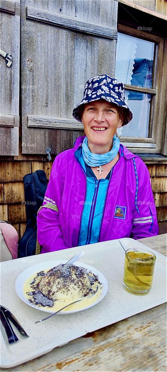 Ecstatically smiling our lady is about to consume a “Germknödel”, a Bavarian delicacy at the “Münchner Haus”, “Munich’s House” inn at 2950 m: a spiced plum jam filled yeast dumpling topped with butter and poppy seeds. 2024. Hypnotic Productions