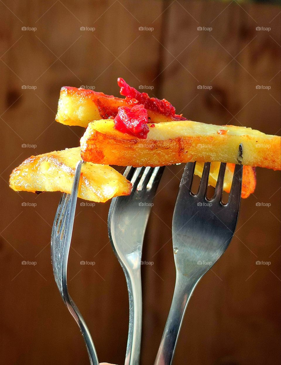 Fried potato slices are strung on three metal forks.  There is ketchup on the potato