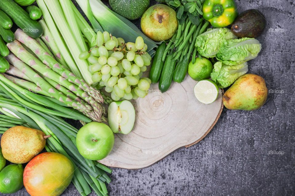 Set of green vegetables and fruits from green onions, celery, asparagus, wheatgrass, green beans, peppers, zucchini, cucumbers, green lettuce, mint, pears, grapes, mango, avocado, apples and limes with a wooden log cabin on a dark stone background, f