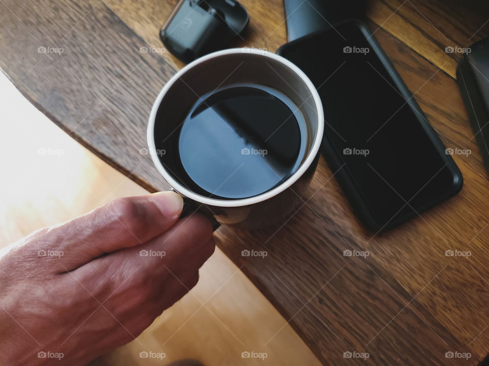 Favorite coffee mug flatlay. Man placing his favorite coffee mug down on an oak antique wood surface with his cell phone on the right and his wireless ear buds on the left.