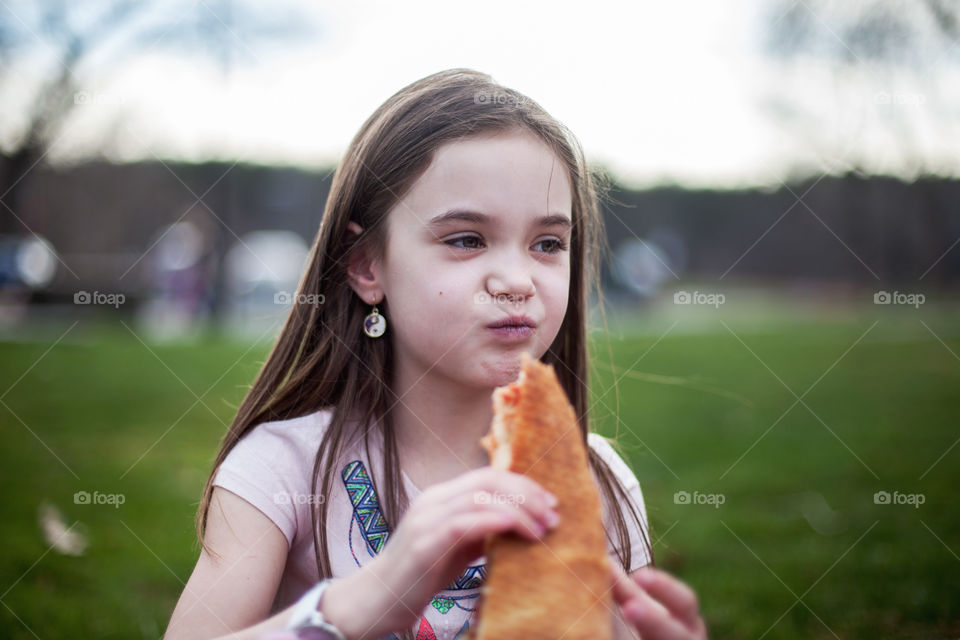 Cute, little brunette girl eating pizza outside on the grass in summer with a smirk on her face. 