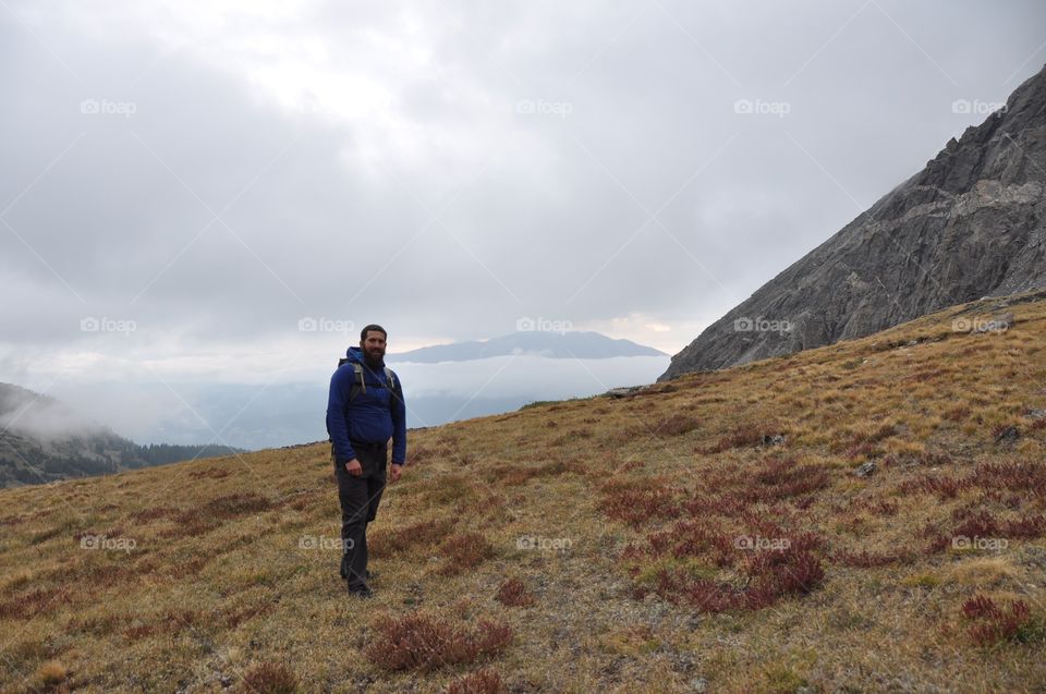 A man stands alone on an alpine tundra with mountains and clouds in the background. Adventure awaits!