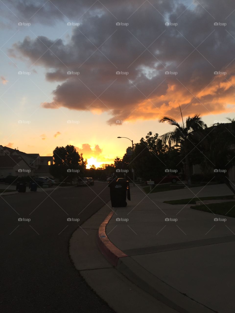Sunset with clouds and sky. Silhouette of buildings, trees, lamppost, and road. 