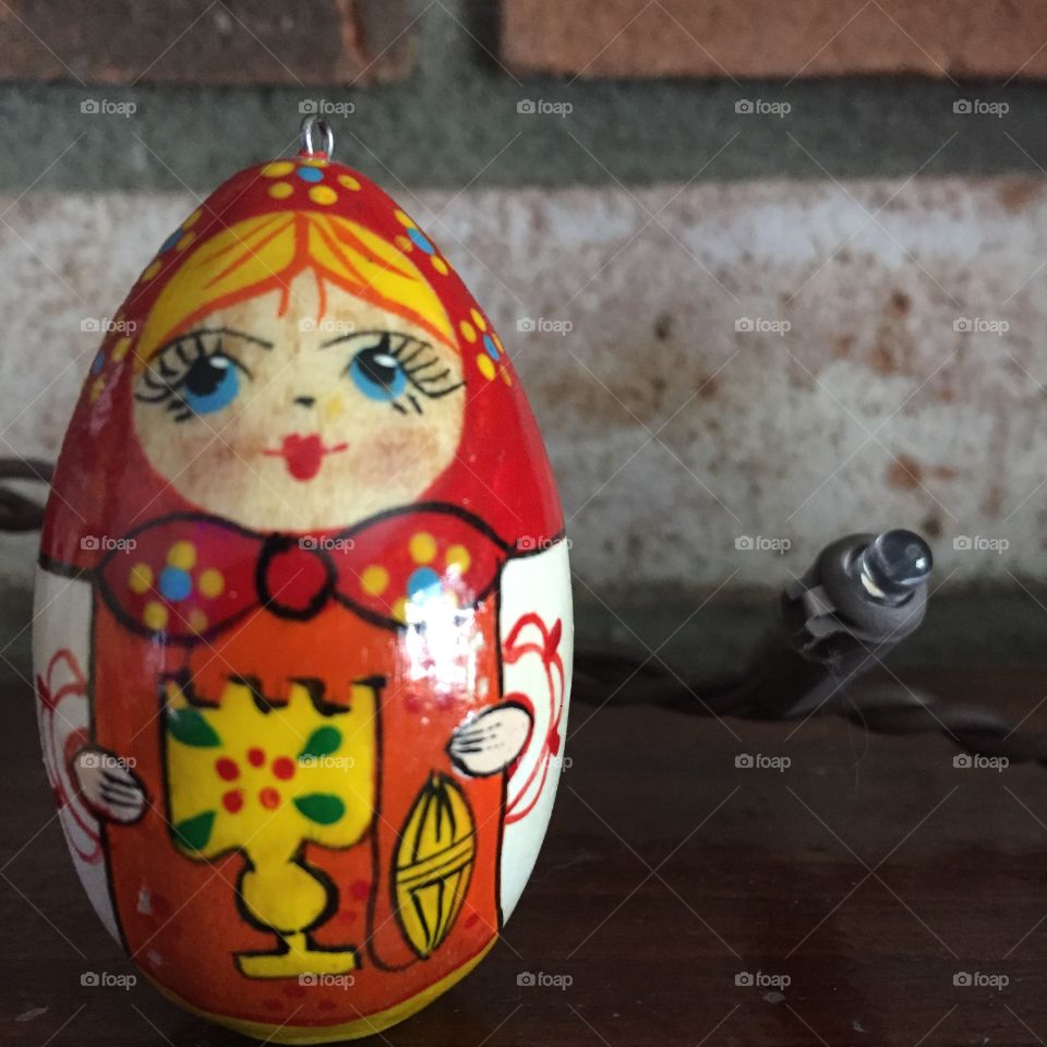 A Christmas ornament that represents a Russian nesting doll. It rests next to an unlit LED bulb in front of a brick wall.