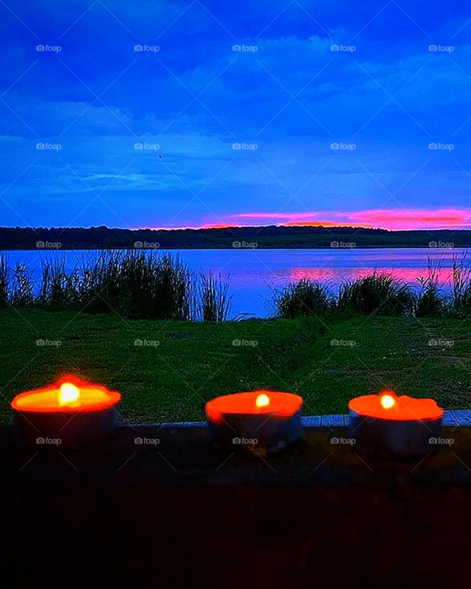 Village or city. River spaces. Evening. Three candles are burning in the foreground. In the darkness, a bank of green grass is visible, the expanse of blue water from which a purple sunset is reflected. Across the river you can see a small field