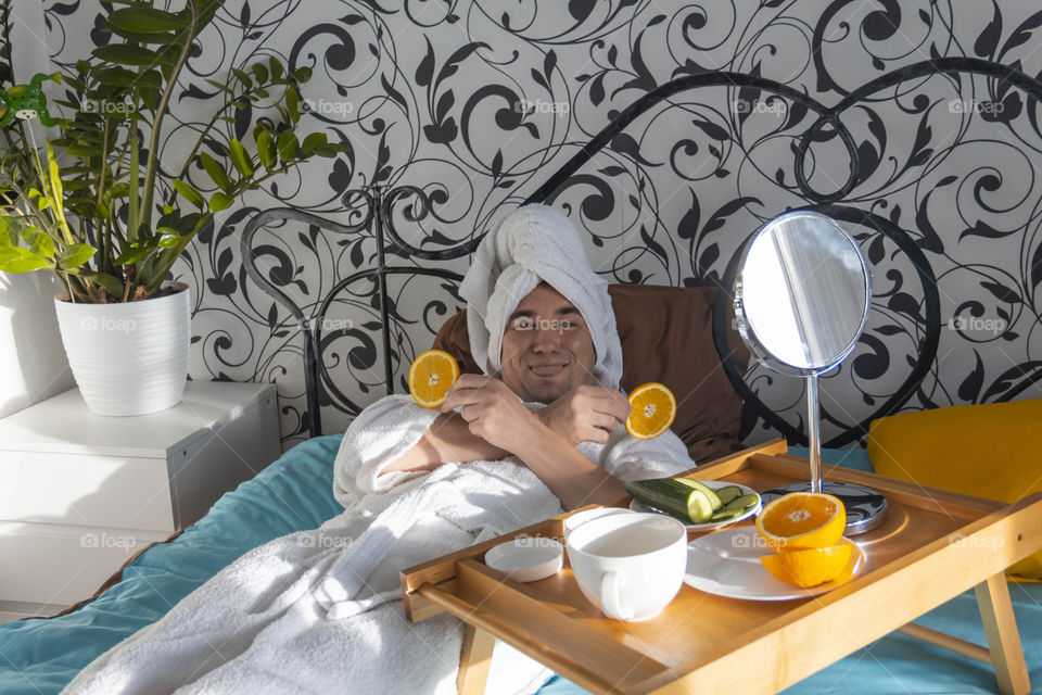 A man and home spa care at home takes care of himself and smears his face with cream, does a manicure at home on a blue background and with oranges and rests.