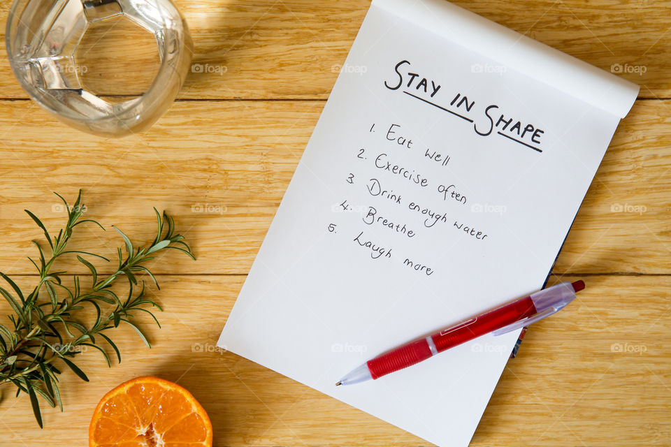 Staying in good shape handwritten list of things to keep you on track to live healthy physically and mentally. Lay flat on wooden background with glass of water with notepad pen citrus fruit and herbs.