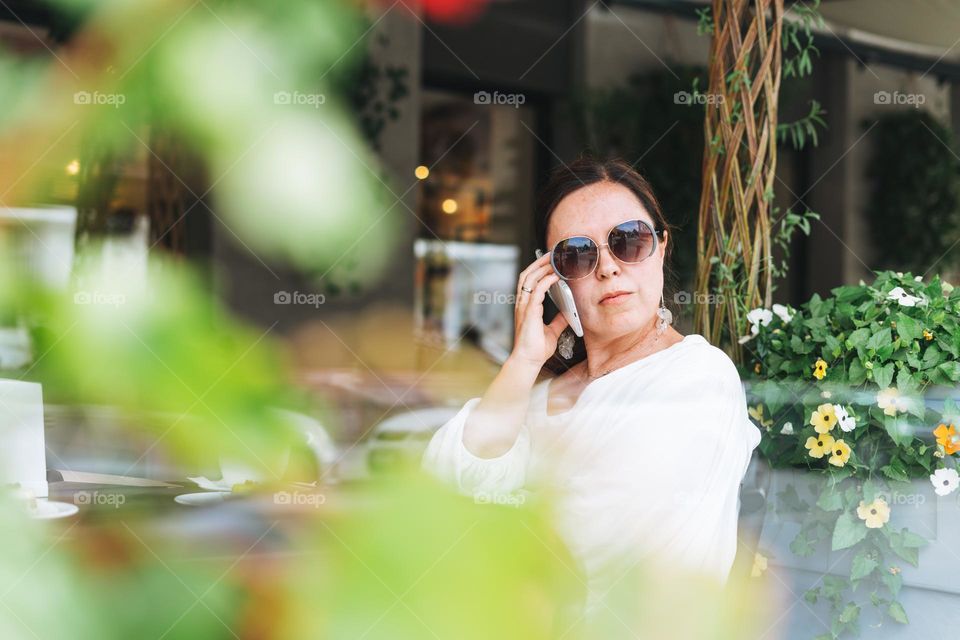 Portrait of smiling beautiful brunette middle aged woman in sunglasses in white clothes with coffee at terrace of the summer cafe