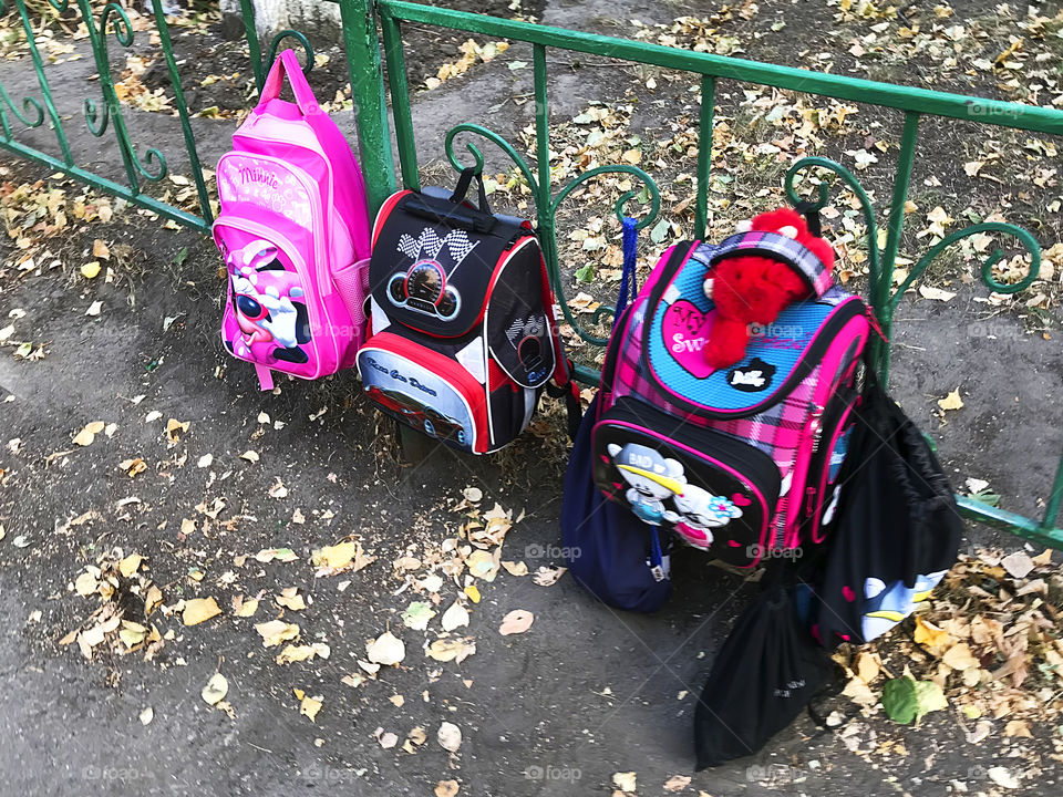 Colorful backpacks of students hanging on the fence above the ground with autumn fallen leaves 