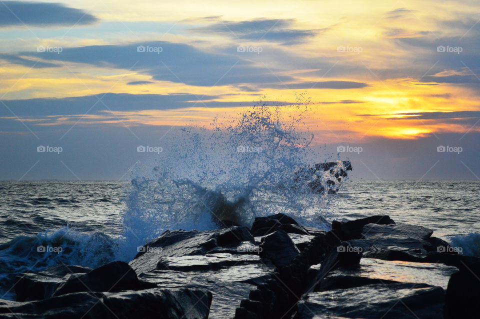 A wave crashes on rocks of a beach shore as the sunsets 