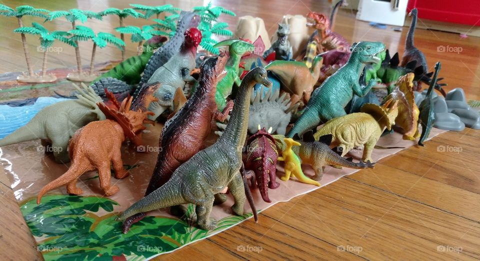 Kids toy colorful dinosaurs arranged on a pretend play mat against a hardwood floor.