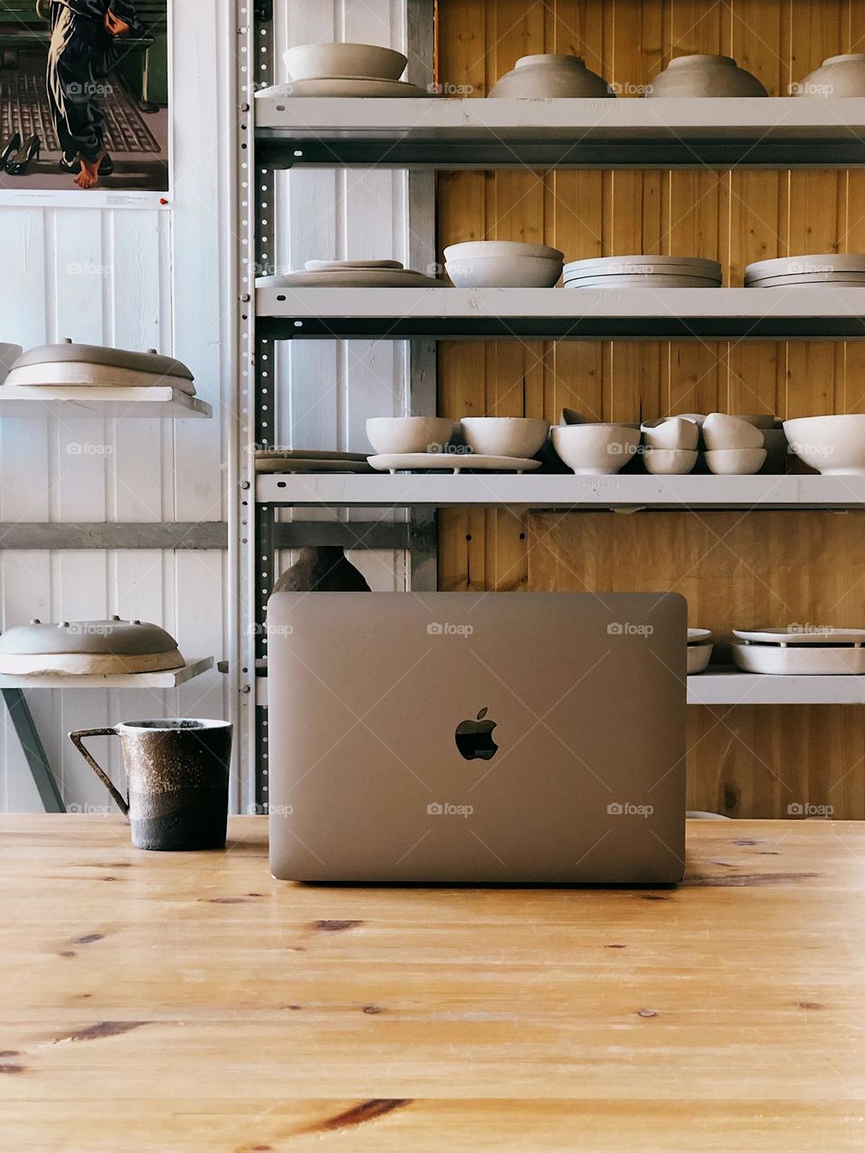 An open computer stands on a wooden table by a window in ceramic studio, nobody 