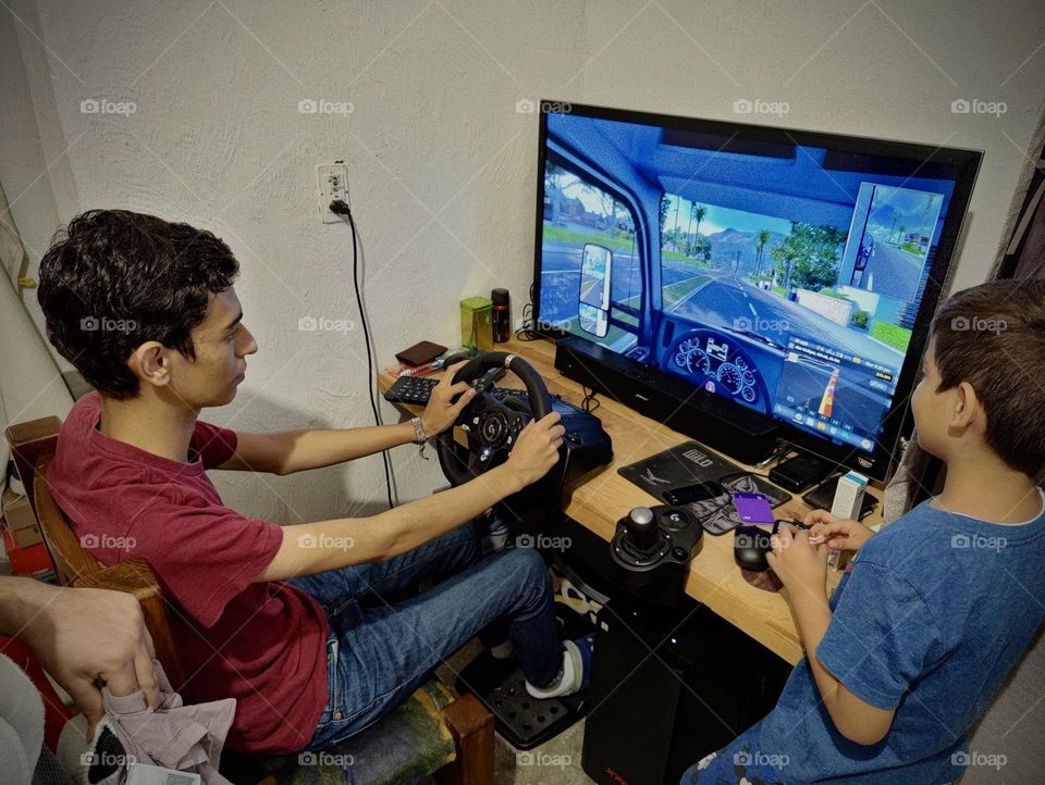 Boy sitting in a chair, playing a driving video game, while his brother looks him playing.