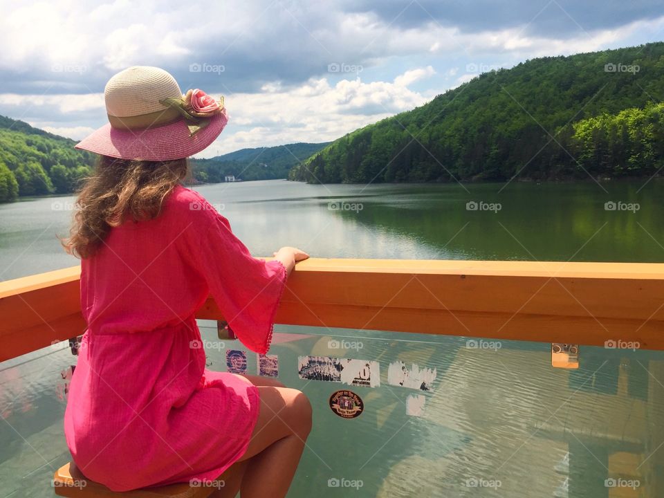 Back of woman with pink dress and pink summer hat looking at the lake on pontoon