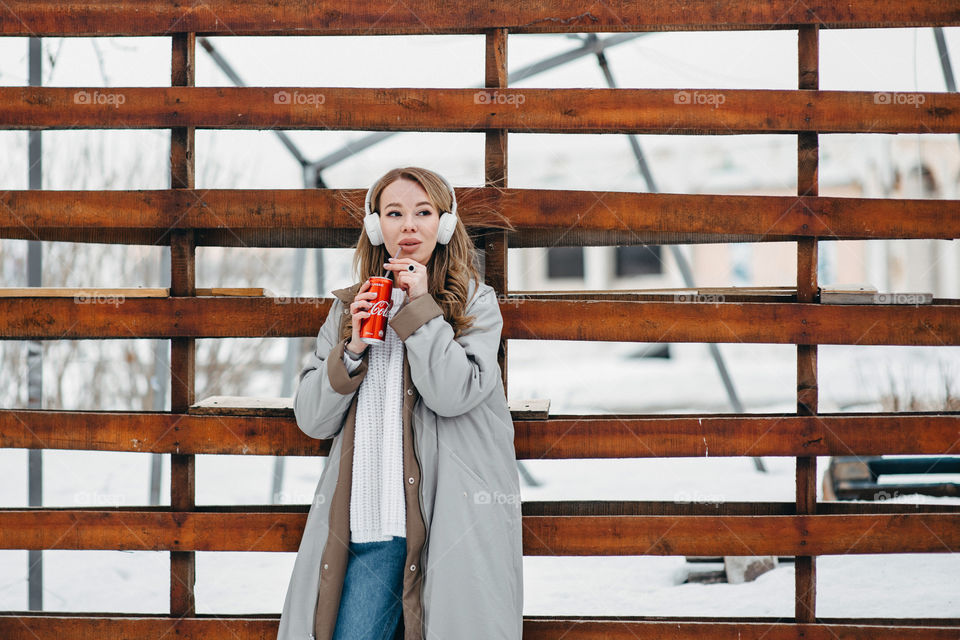 A woman in white headphones listening to music and drinking Coca Cola. Winter cold season. Grey coat. Stylish winter outfit