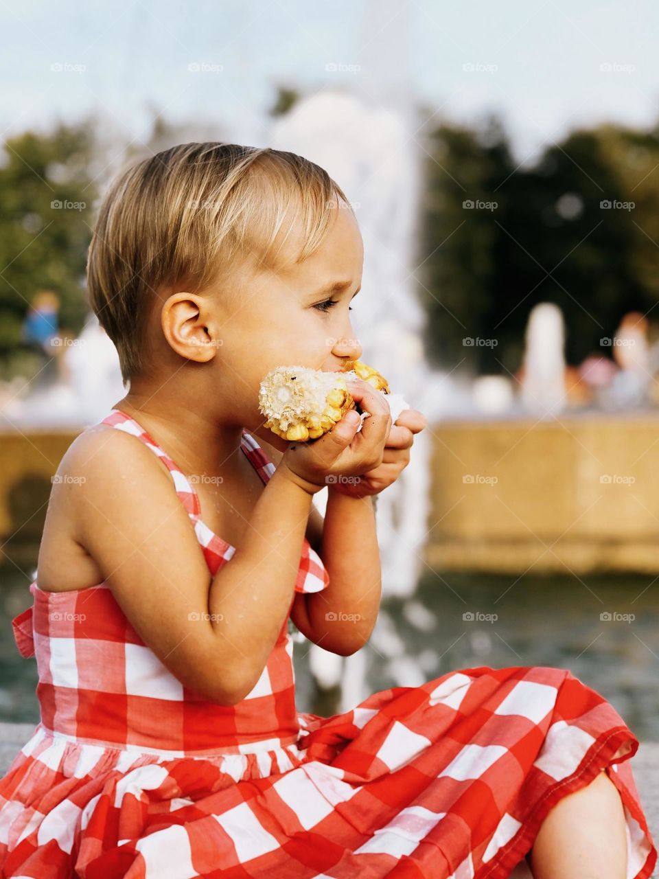 Cute little girl with short blonde hair wearing red dress sitting near fountains and eating corn, portrait of child 