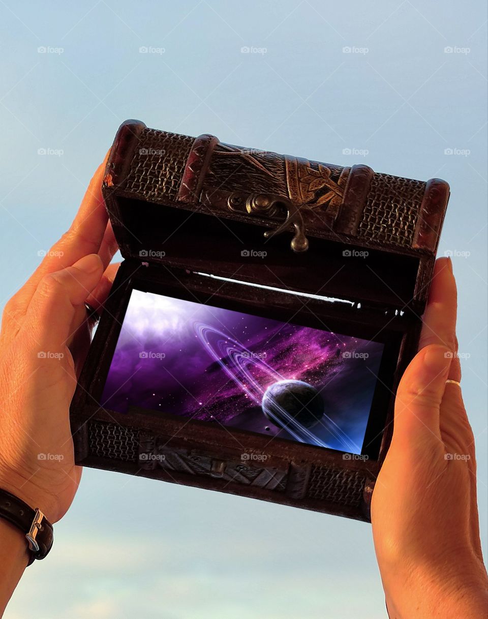 Cosmotogoria. Women's hands hold a wooden carved box against a clear blue sky. At the bottom of the box is a purple cosmos variant with a planet and a dust ring around it. All shades of purple