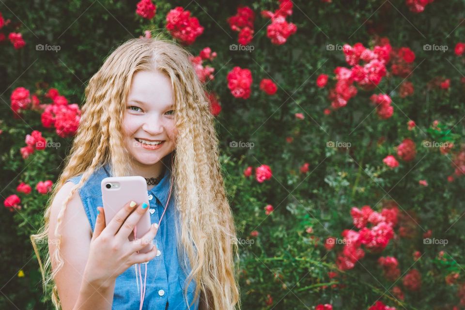 Natural Long hair blond curly hair teen girl smiling looking at camera holding phone against pink roses backpack 