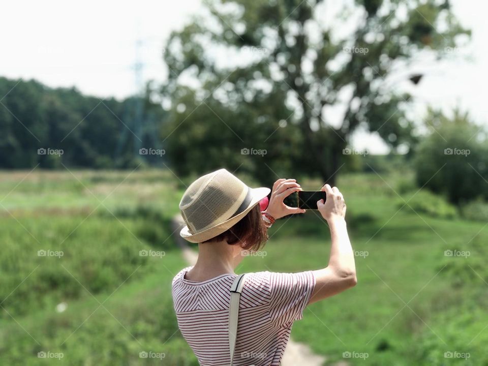 Portrait of a woman in a hat taking photo on smartphone, summer landscape 