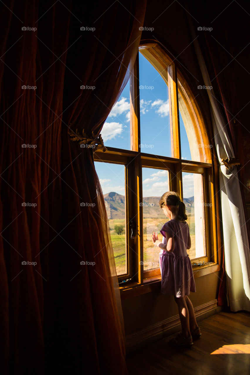 Love the natural light of this image. Photo of a girl standing by large window looking out.