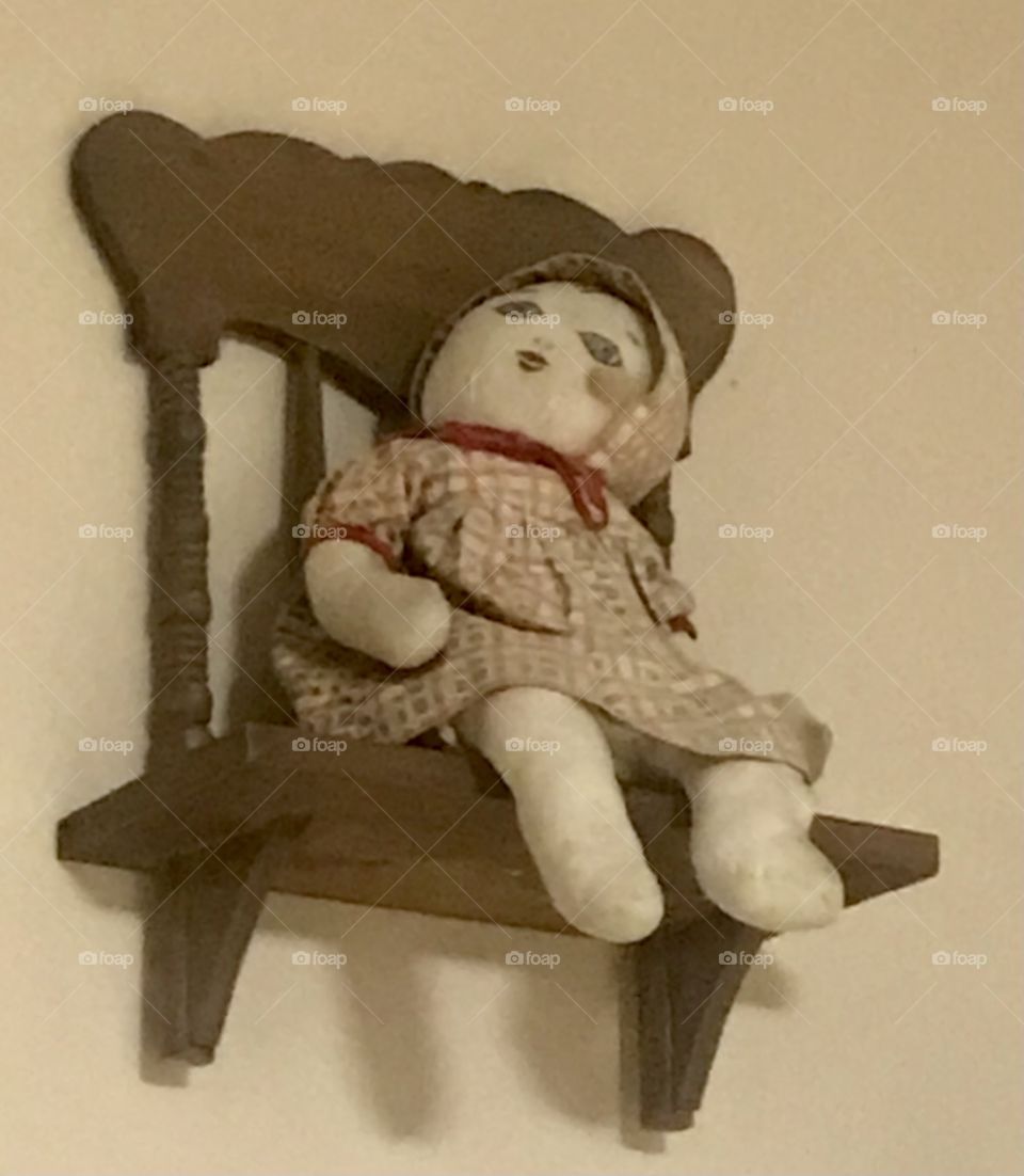 An old rag doll sitting on a wooden chair that hangs on a wall 