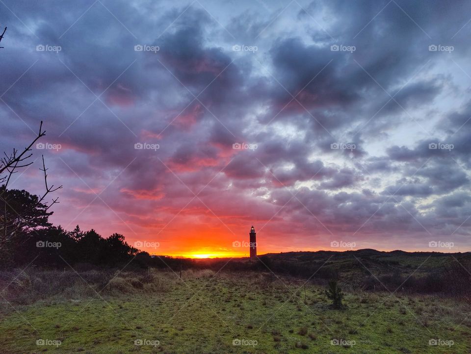 Colorful red, orange, yellow sunset with blue and purple clouds at the dunes with lighthouse in background.