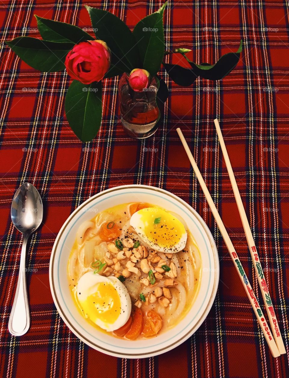 A nice table setting of an Asian inspired dish placed in a bowl & as the center of the photo. Around it are some chopsticks for a more traditional feel or a spoon for a more American way. Pink flowers placed in a small vase on a plaid table cloth. 