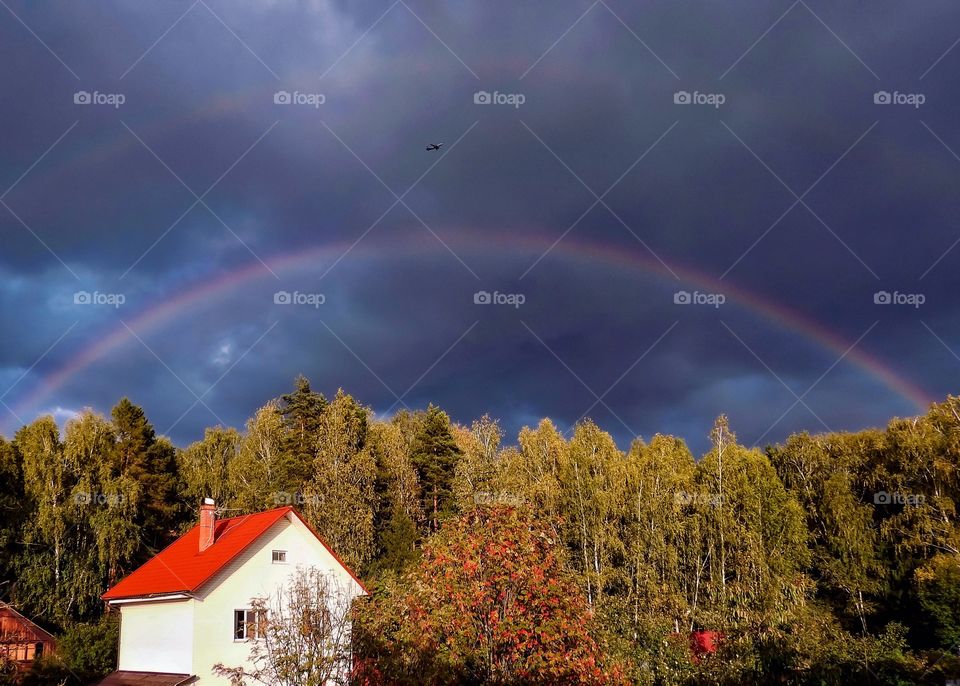 Rainbow over the forest and the house against the background of dark clouds