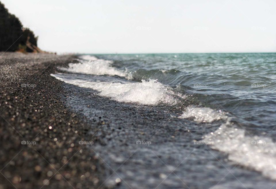 Turquoise waves with foam of a salt lake running onto a black pebble beach along a cliff cliff